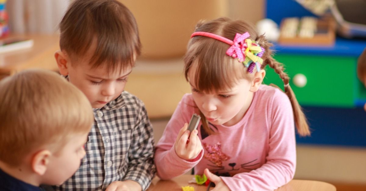 Three young children sit at a small table, examining toy blocks while a girl holds a piece close to her face.