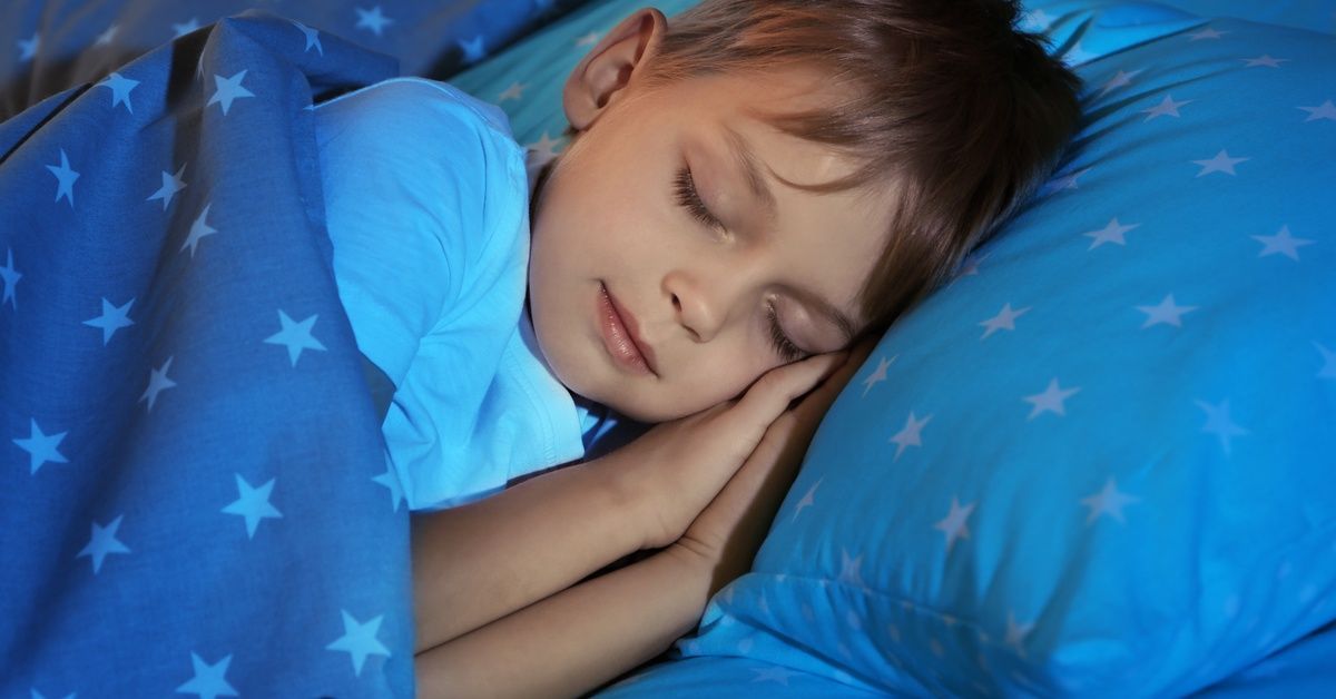 A young child sleeps peacefully on a pillow under a star-patterned blanket with his hands tucked near his face.