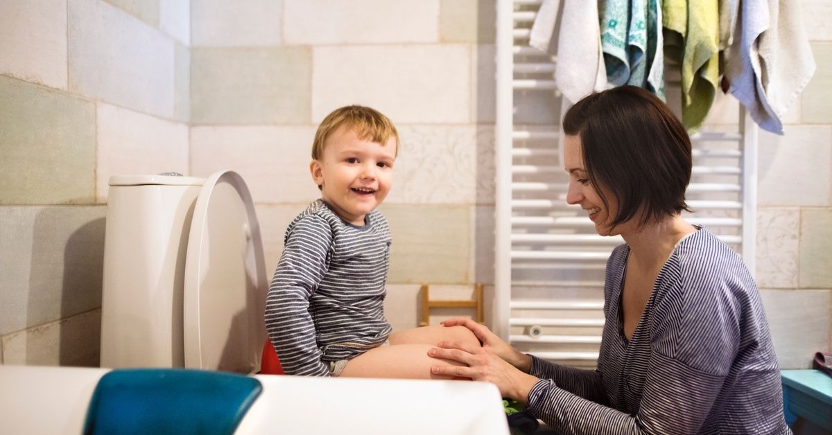 A young child sits on a toilet while their parent kneels nearby, smiling and offering support in a bright bathroom.