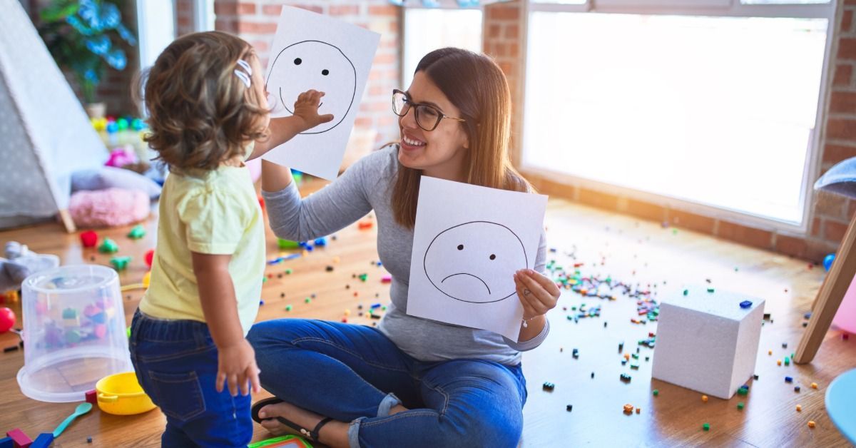 A therapist sits on the floor and shows a child emotion cards with happy and sad faces in a bright playroom.