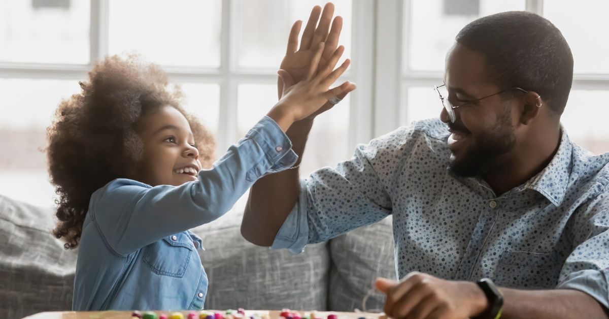 A smiling father gives a high five to a young girl at a table with colorful beads. A large window lets in light behind them.