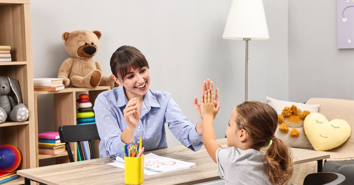 A smiling therapist gives a high five to a girl at a table. A shelf with crayons, toys, and a teddy bear is behind them.