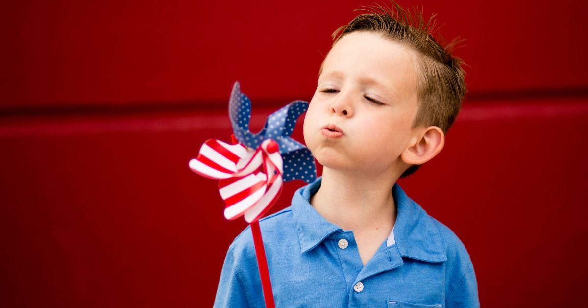 A young boy blows on a red, white, and blue pinwheel while standing in front of a bright red background.