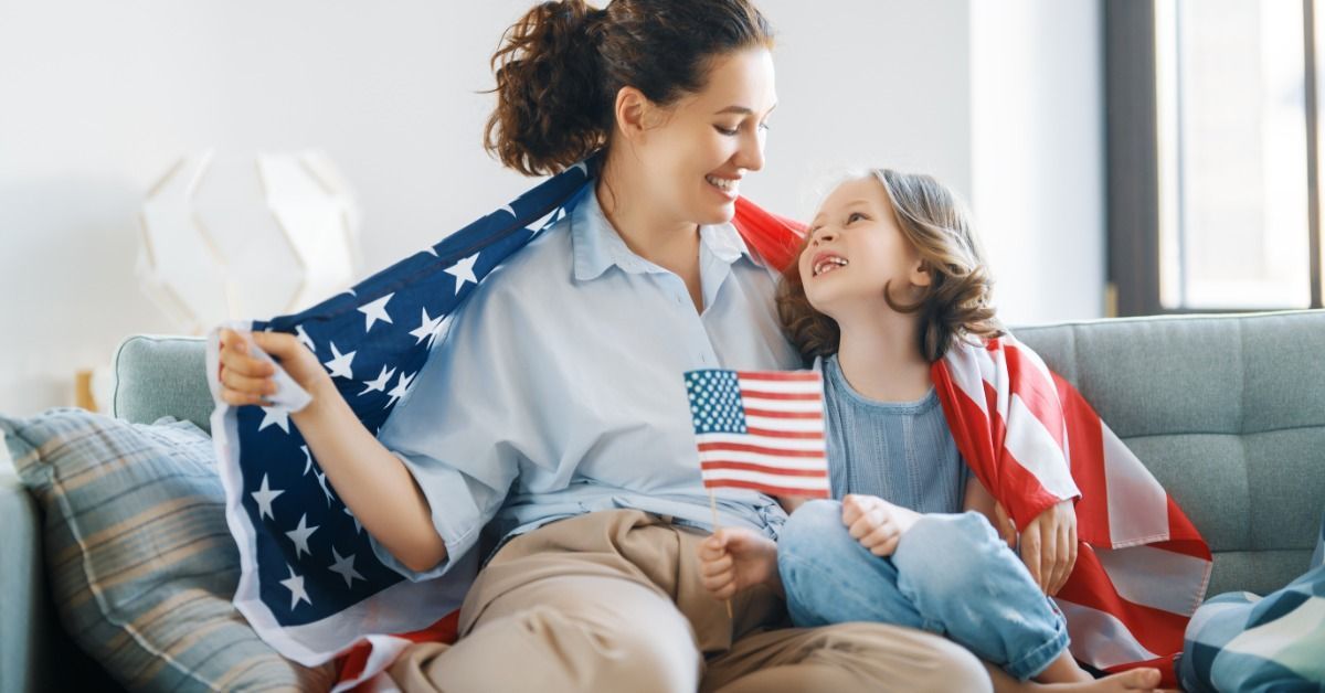 A mother and a young child wrapped in an American flag sit on a couch. The child holds a smaller flag in their hand.