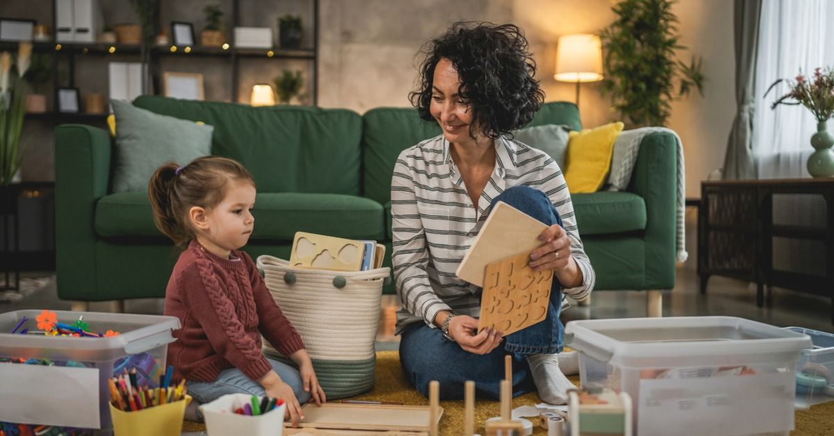 A mother sits on the floor with a young child and holds a wooden puzzle while they put toys away in a cozy living room.