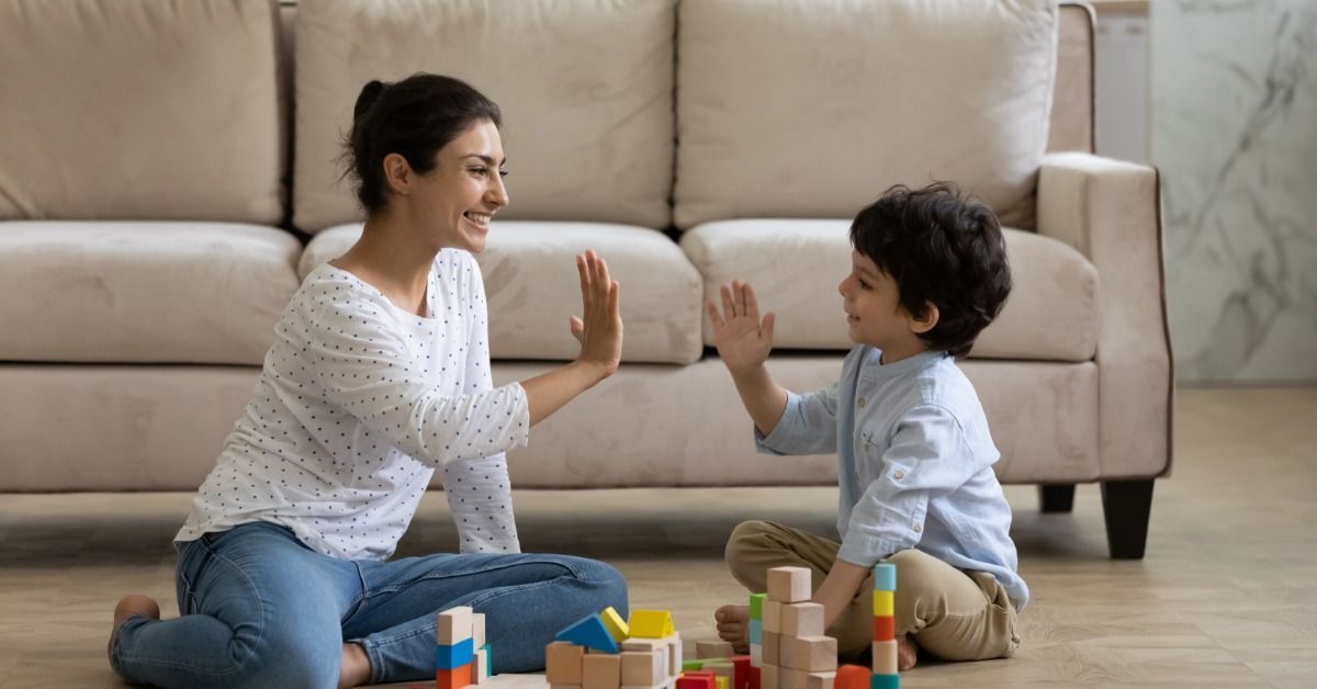 A mother and a young child sit on the floor and give a high five to each other while playing with colorful wooden blocks.