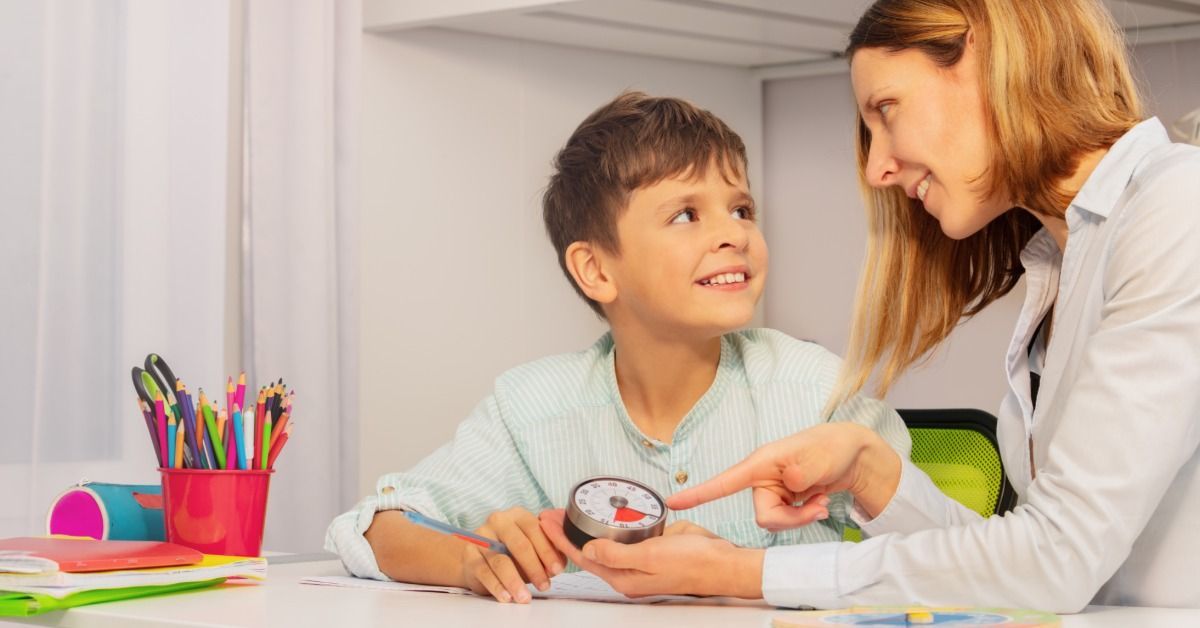 A therapist points to a timer while smiling at a young boy seated beside pencils and school supplies.
