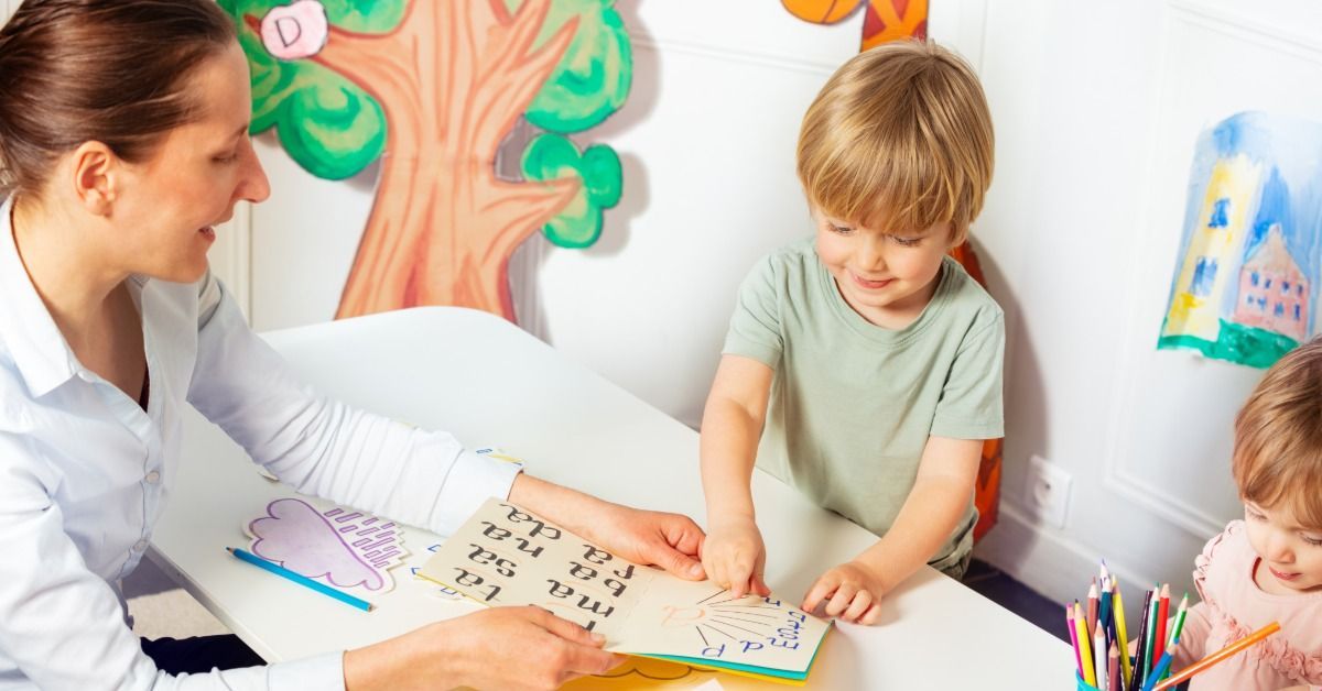 A therapist helps two young children practice reading and matching activities at a small classroom table.