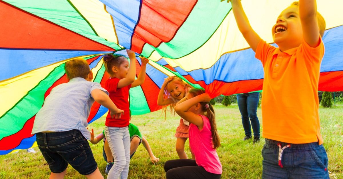 Children lift a colorful parachute during a lively outdoor summer camp game on a sunny grassy field.