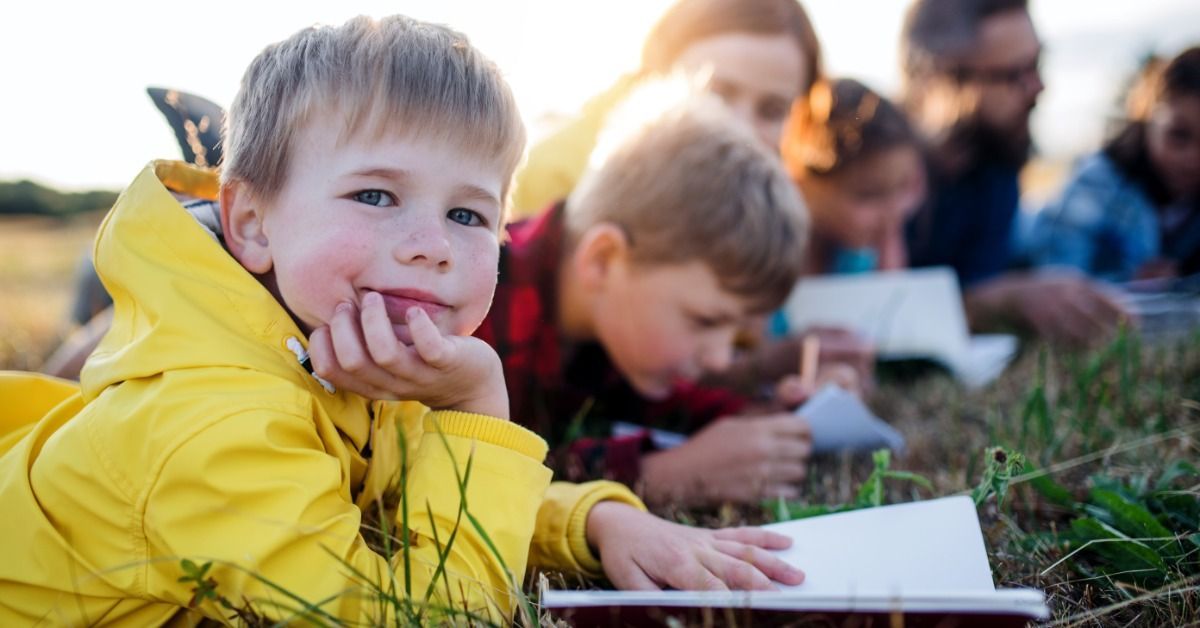 A young boy in a yellow jacket looks at the camera while children draw in notebooks nearby during a nature activity.