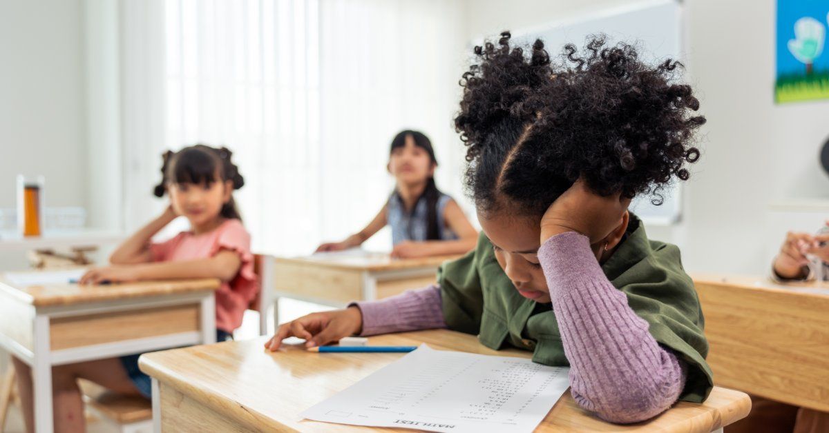 A young student leans over a worksheet at a classroom desk while other children sit quietly behind her.