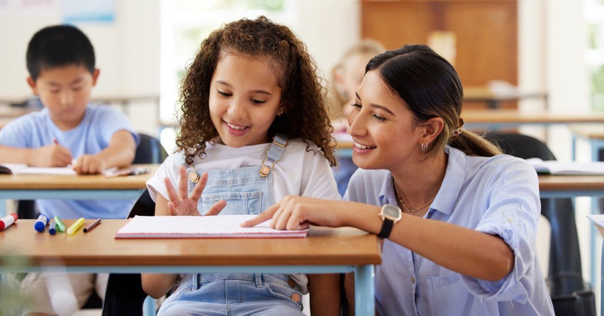 A teacher points to a notebook as a smiling young girl counts on her fingers at a classroom desk.