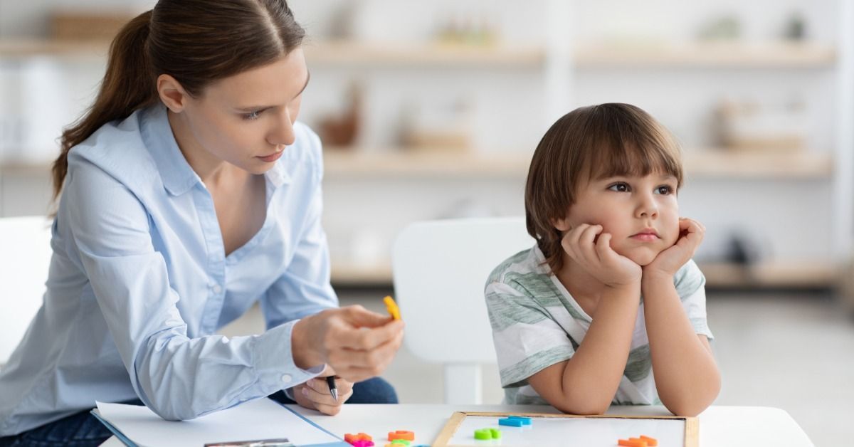 A therapist holds a learning piece while a young child sits at a table with educational shapes during an ABA session.