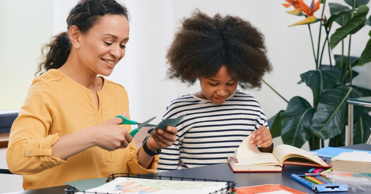 A therapist guides a smiling child through a craft activity while books and classroom supplies sit on a table.