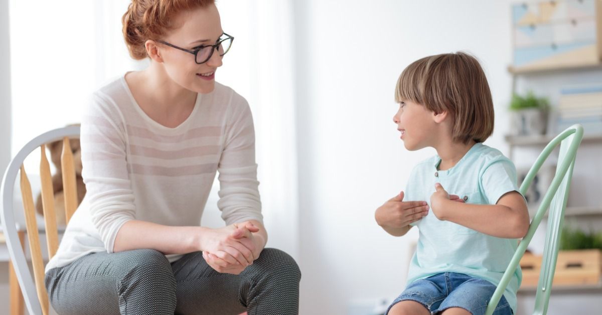 A therapist listens warmly as a young child gestures while sitting on a green chair in a bright therapy room.