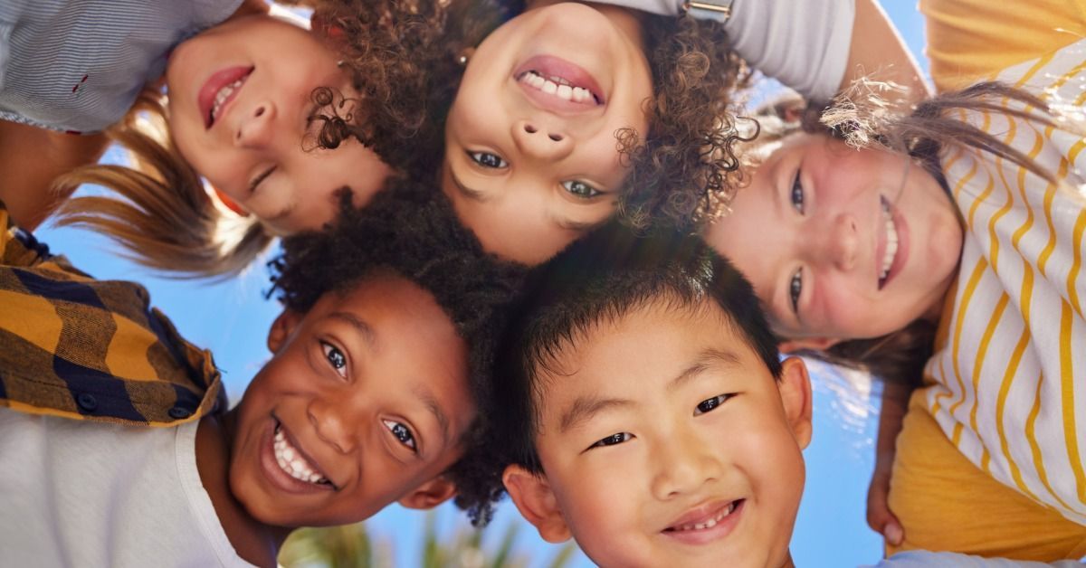 Five smiling children lean into a circle under the bright blue sky and look down toward the camera.