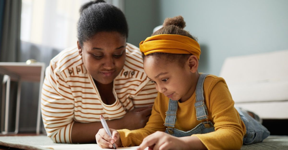 A smiling mother watches her daughter draw with a pencil on the floor in a cozy room filled with soft daylight.