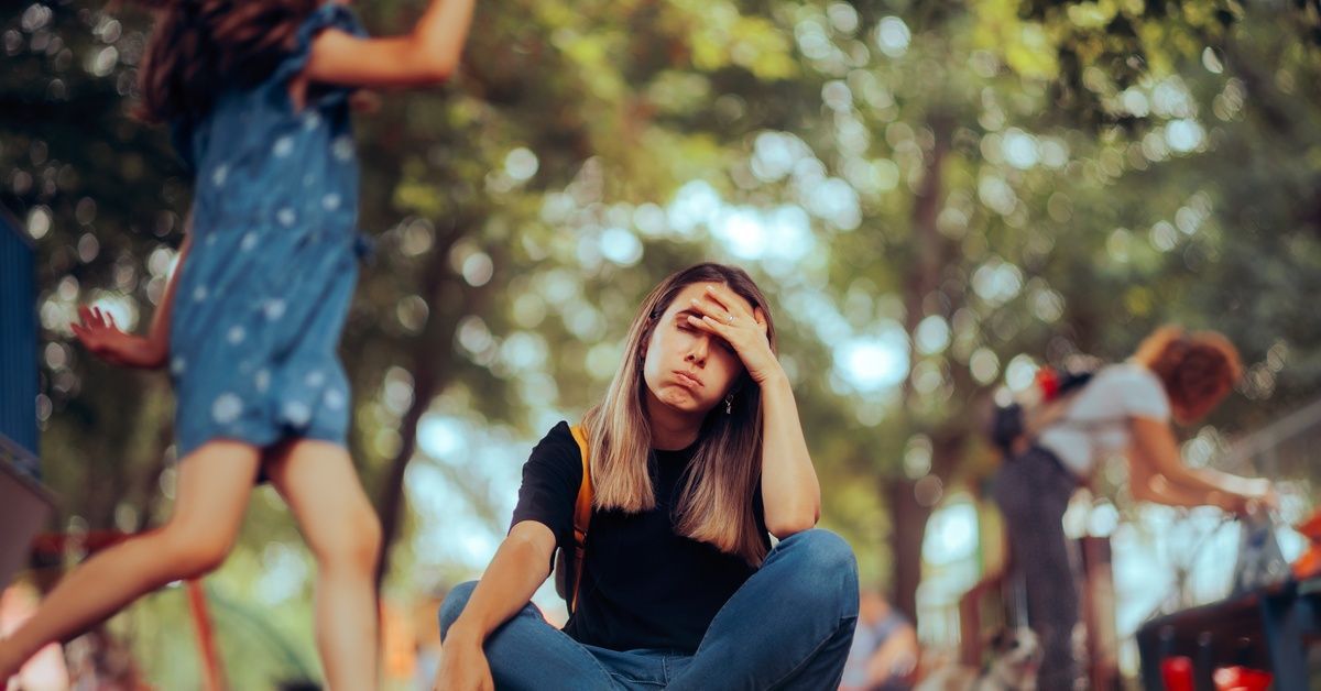 A tired mother sits on the ground at a playground while children run and play around her.