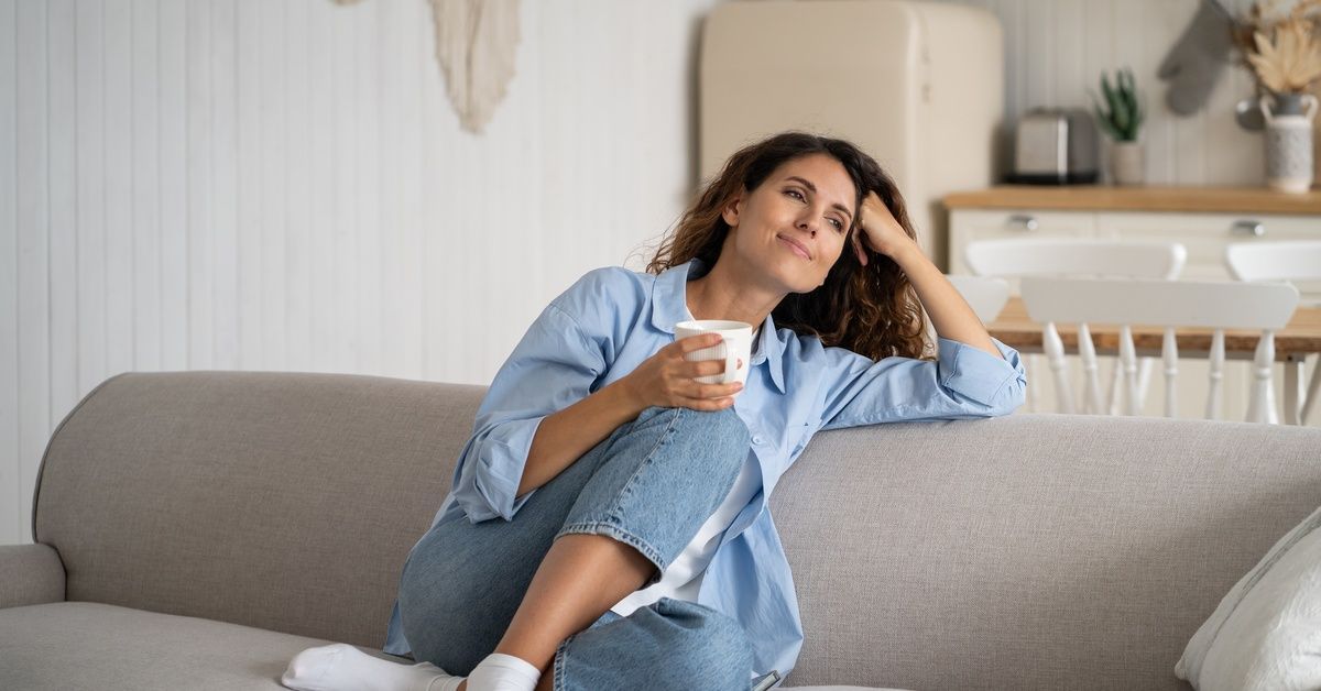 A mother sits on a couch with a mug and takes a quiet moment in a bright, calm living room in her home.