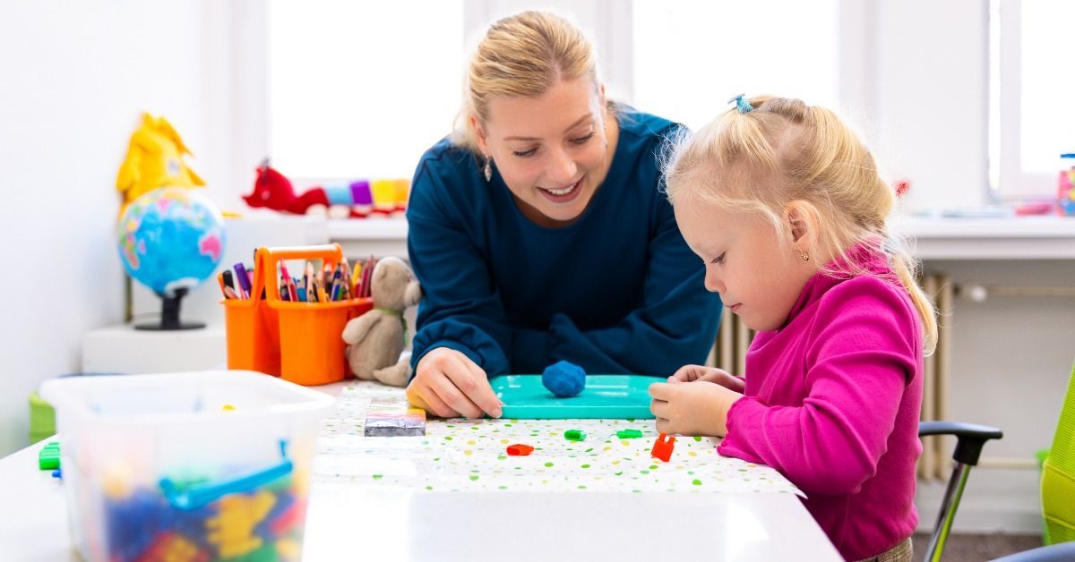 A therapist helps a young girl shape colorful clay at a table surrounded by art supplies in a bright