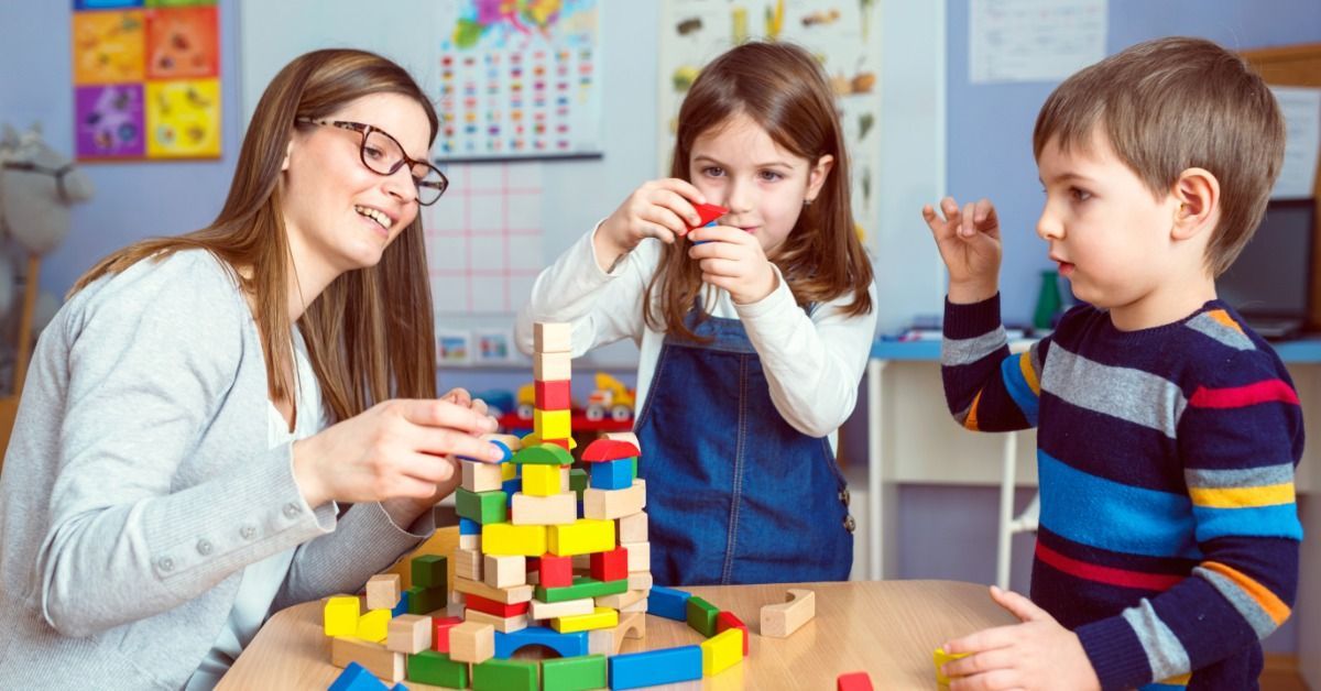 A teacher builds a colorful block tower with two children as they take turns adding pieces at a classroom table.