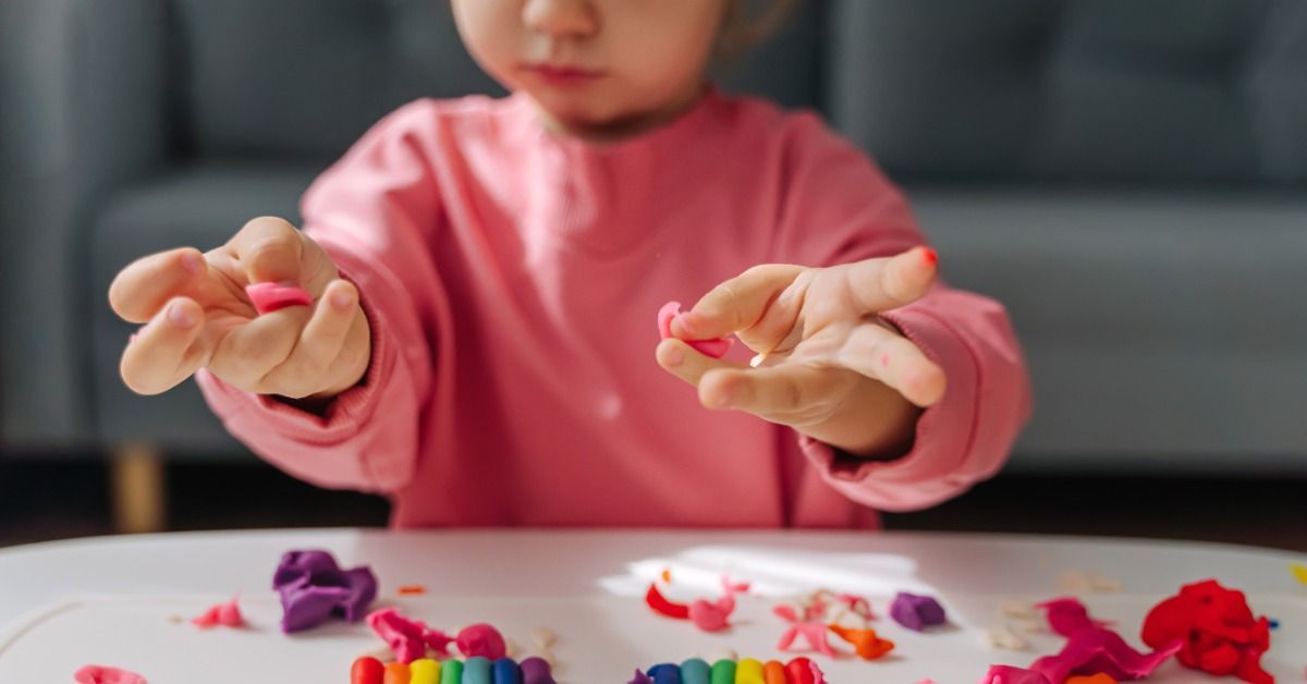 A young child shapes pink clay with both hands beside a colorful clay rainbow and scattered clay pieces on a table.
