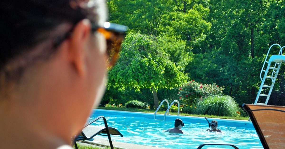 A woman watches two children swim in a backyard pool surrounded by green trees and lounge chairs on a sunny day.