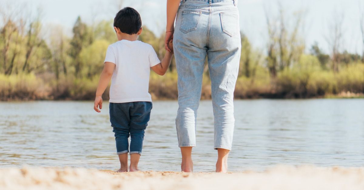 A mother holds her young child’s hand as they walk along a sandy beach beside a calm lake on a bright day.