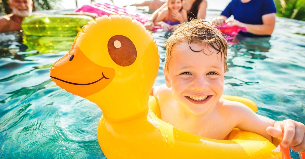 A smiling boy floats in a yellow duck tube as a family watches from the sunny pool behind him on a b