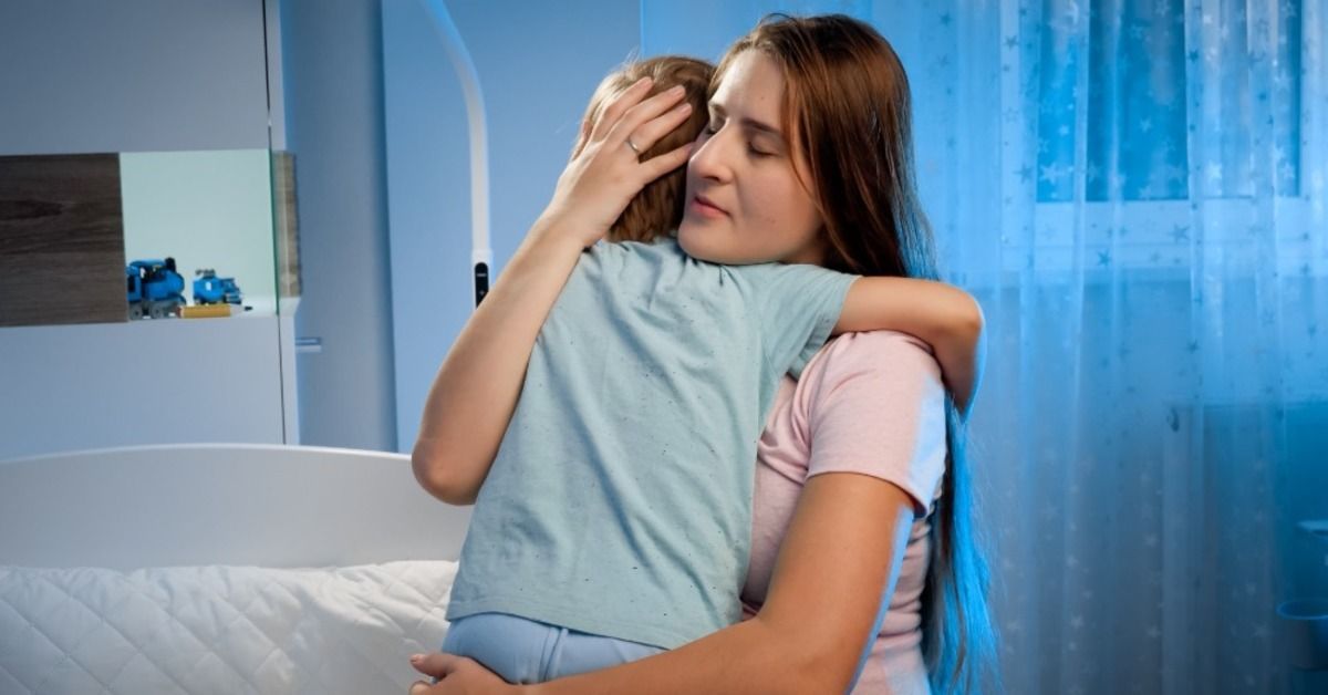 A mother hugs and comforts a child beside a bed as blue evening light fills the softly lit bedroom a