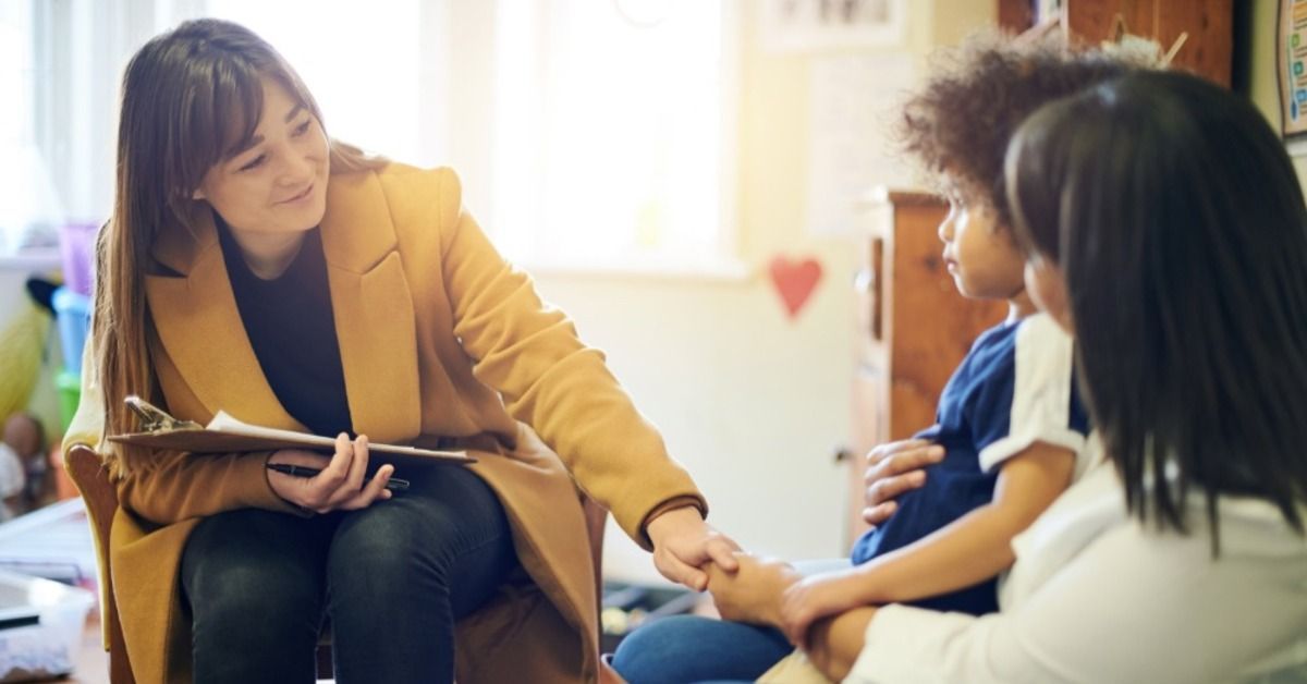 A therapist holds a child's hand while speaking with the child's mother as they all sit together in a bright office.