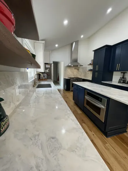 Modern kitchen with white marble counters, dark blue cabinets, stainless appliances, and wood floor under recessed lights