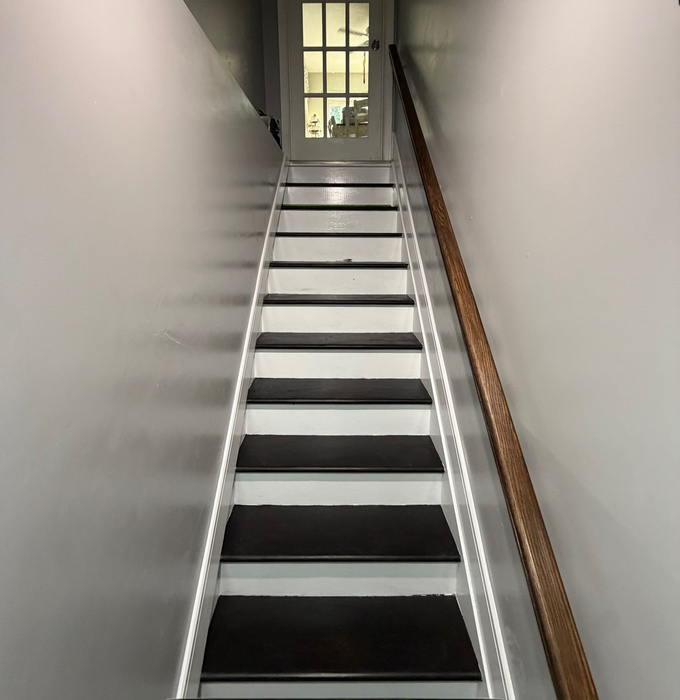 Narrow staircase with white walls, dark steps, and a wooden handrail leading to a window.