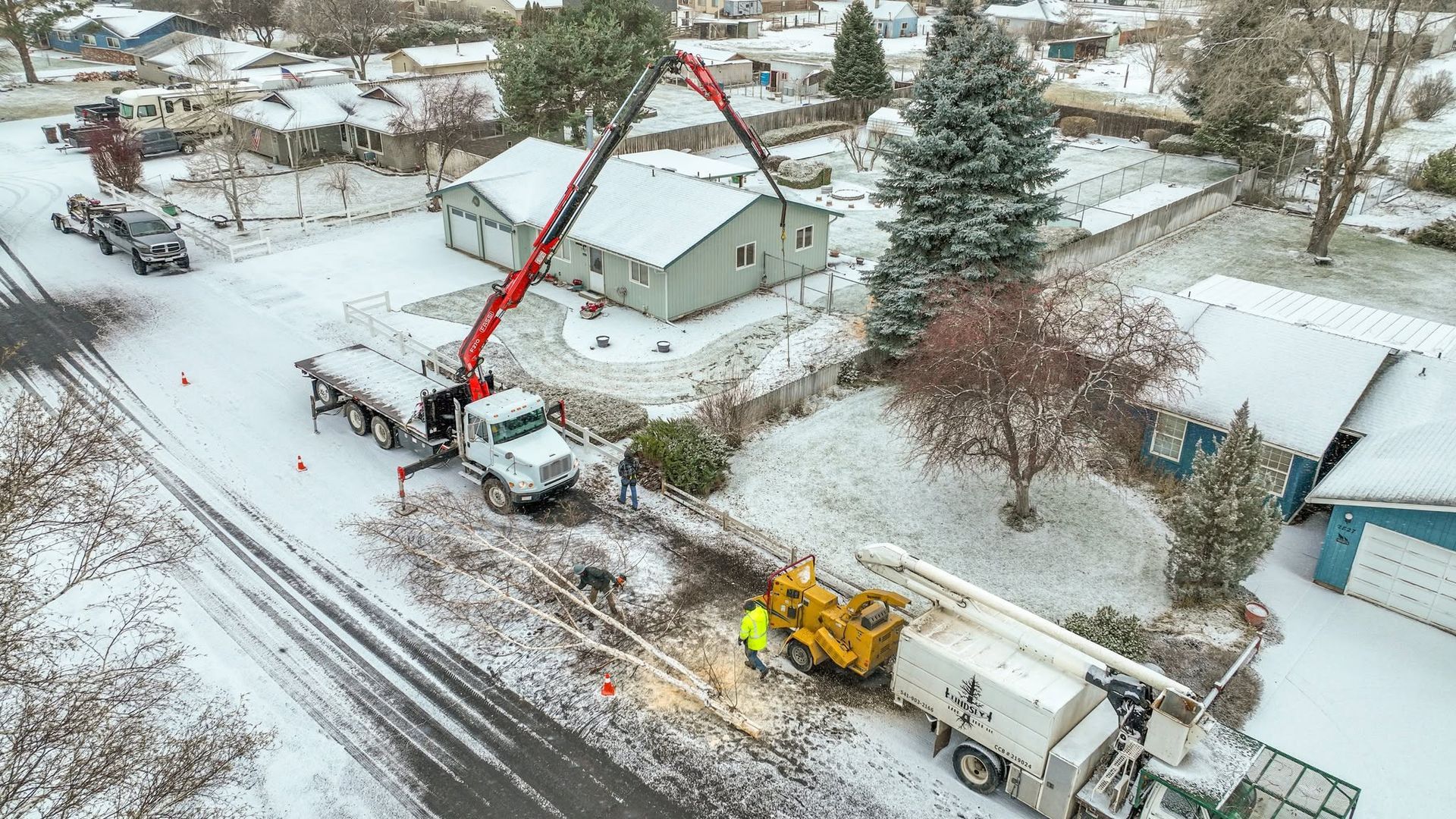 Snowy residential street with a red crane truck and yellow utility truck beside houses and trees