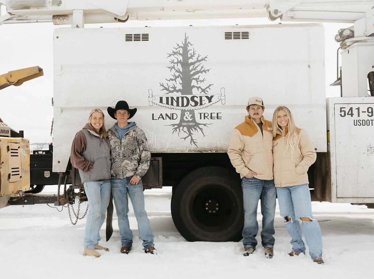 Four people posing in winter clothes beside a snow-covered truck with a tree logo.