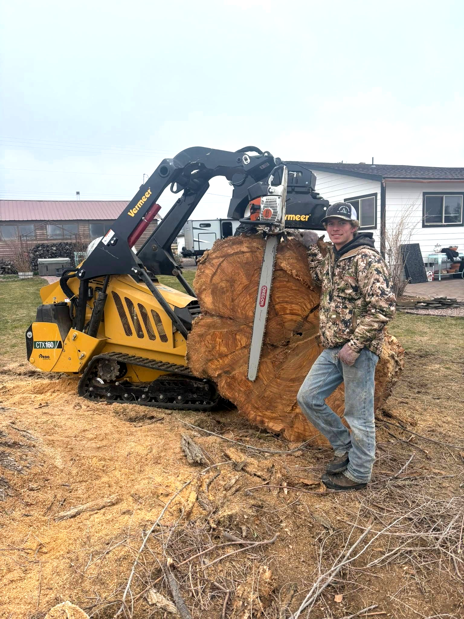 Man standing beside a yellow skid steer lifting a huge round hay bale in a farmyard