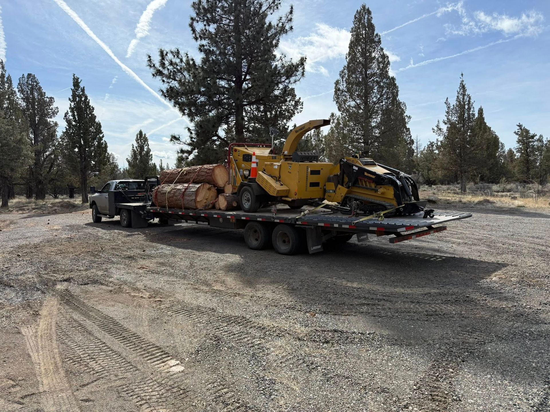 Yellow construction equipment on a flatbed trailer in a gravel lot with trees and blue sky.