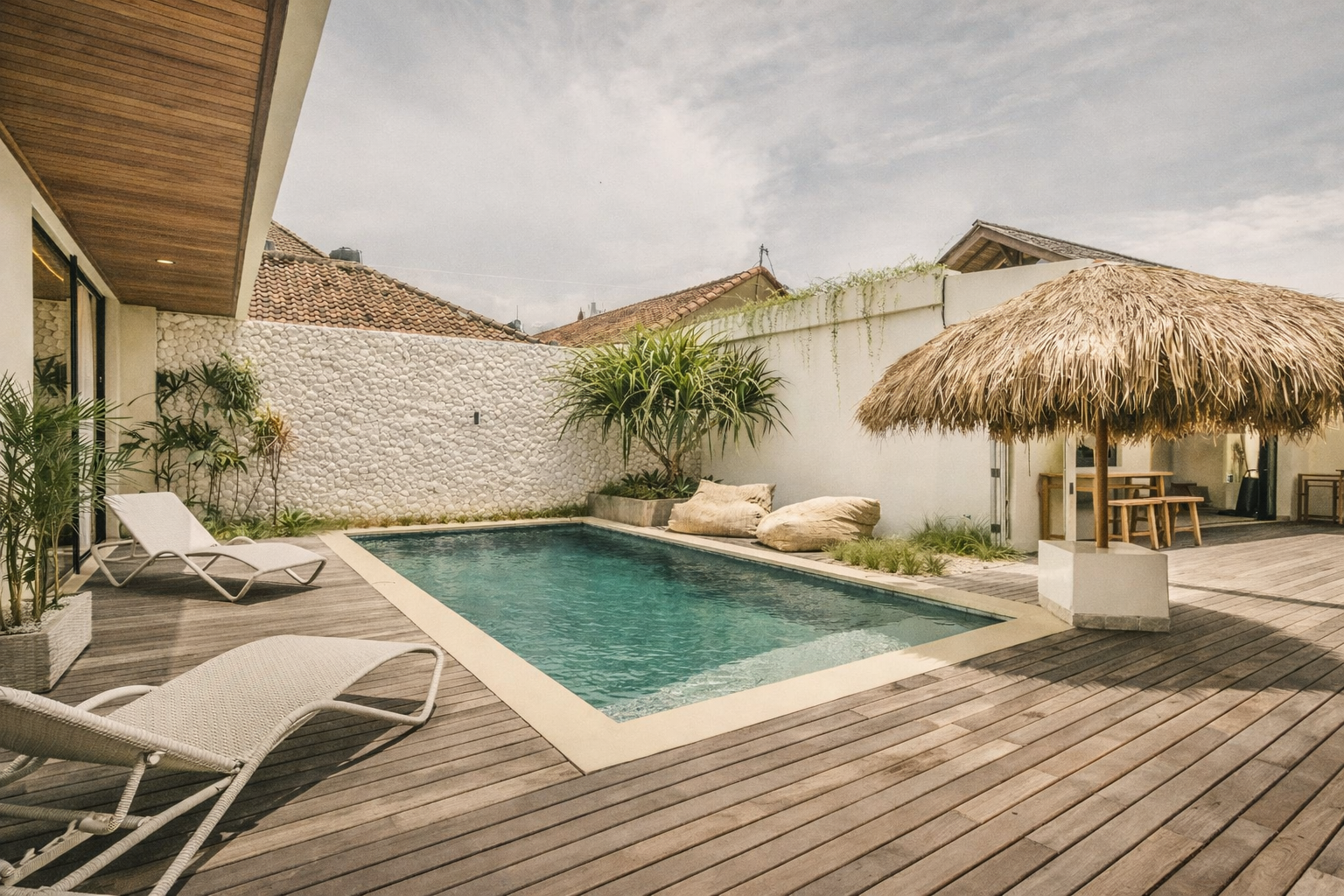 Pool area with lounge chairs, thatched-roof umbrella, and white stone wall.