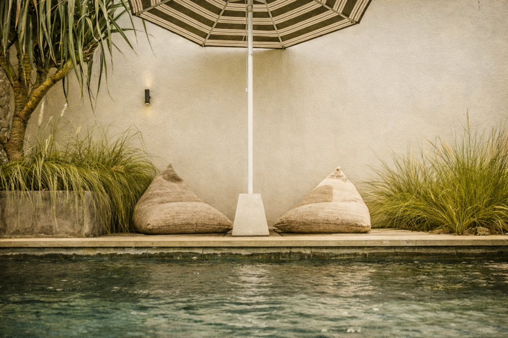 Two beige bean bag chairs under a striped umbrella by a pool.