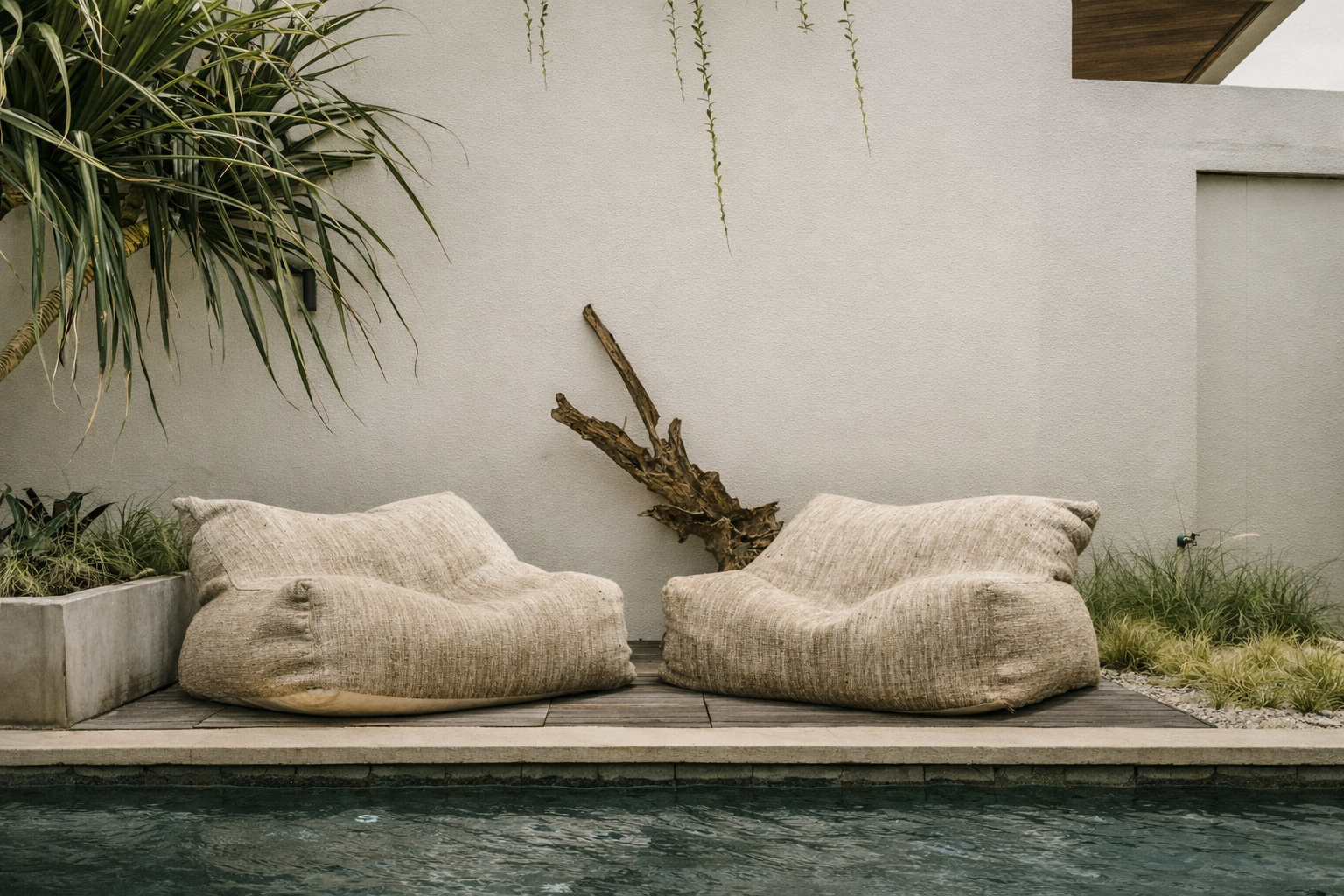 Two tan beanbag chairs sit by a pool against a stucco wall with plants.