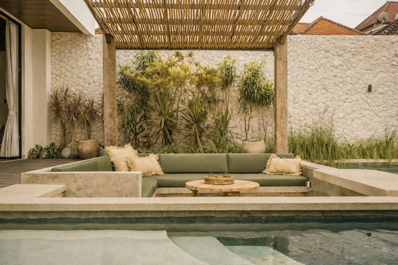 Outdoor seating area with pool, green sofa, woven pergola, and textured white wall.