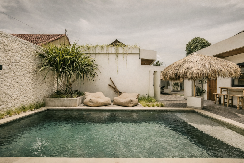 Swimming pool and lounge chairs in a tropical outdoor setting with white walls and thatched roof.