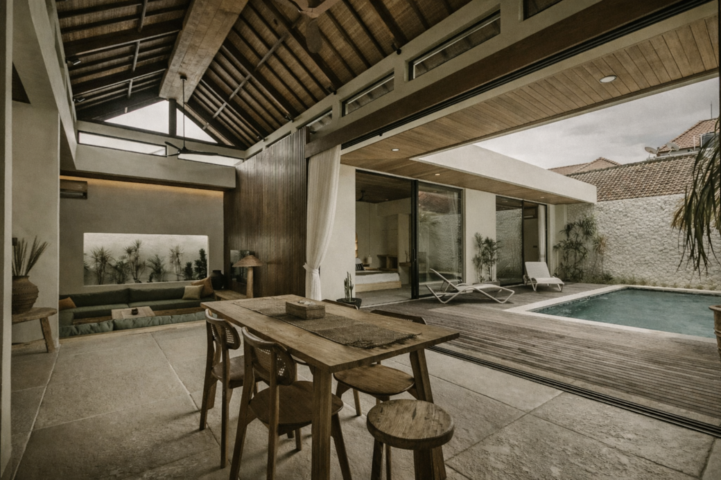 Dining area with wooden table and chairs, opening to a pool and villa.