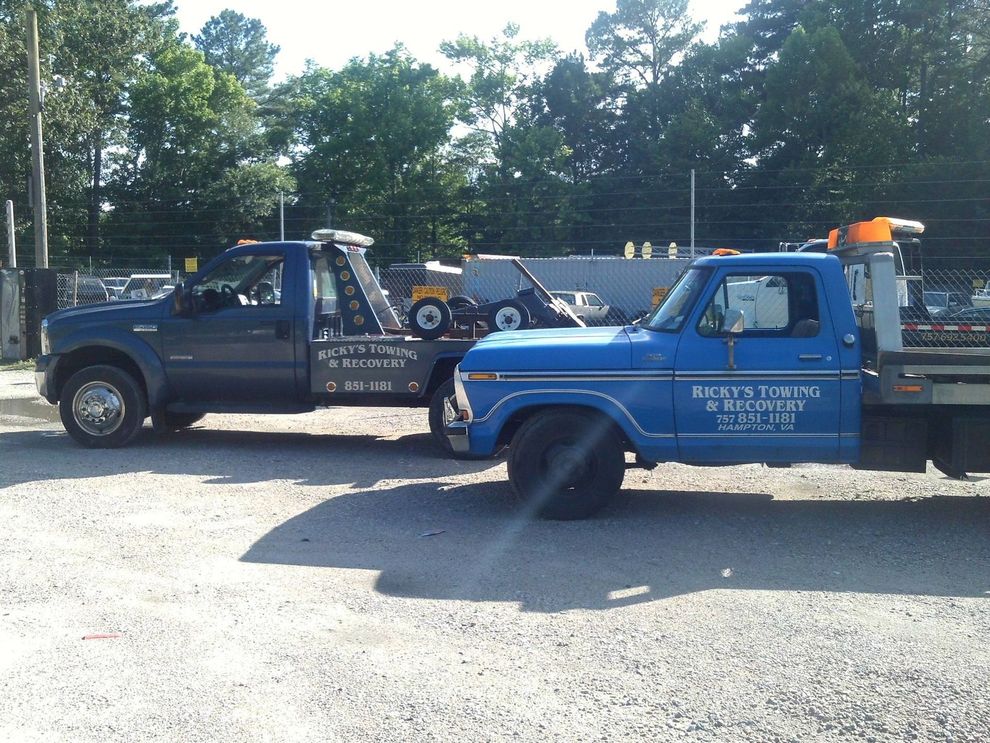Two blue tow trucks parked on a gravel lot under sunny skies.