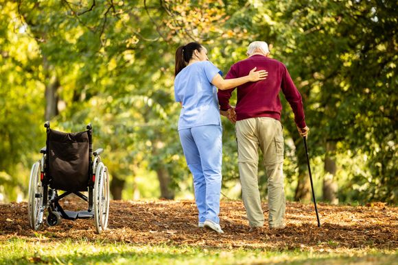 Un operatore sanitario in camice blu assiste una persona che usa un bastone mentre camminano insieme in un parco. Una sedia a rotelle si trova nelle vicinanze.
