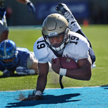 Football player diving into the end zone with the ball, wearing a white uniform and gold helmet on blue turf