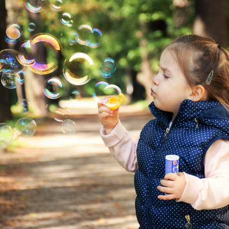 Child blowing bubbles outdoors, holding a bubble wand and bottle in a sunny park