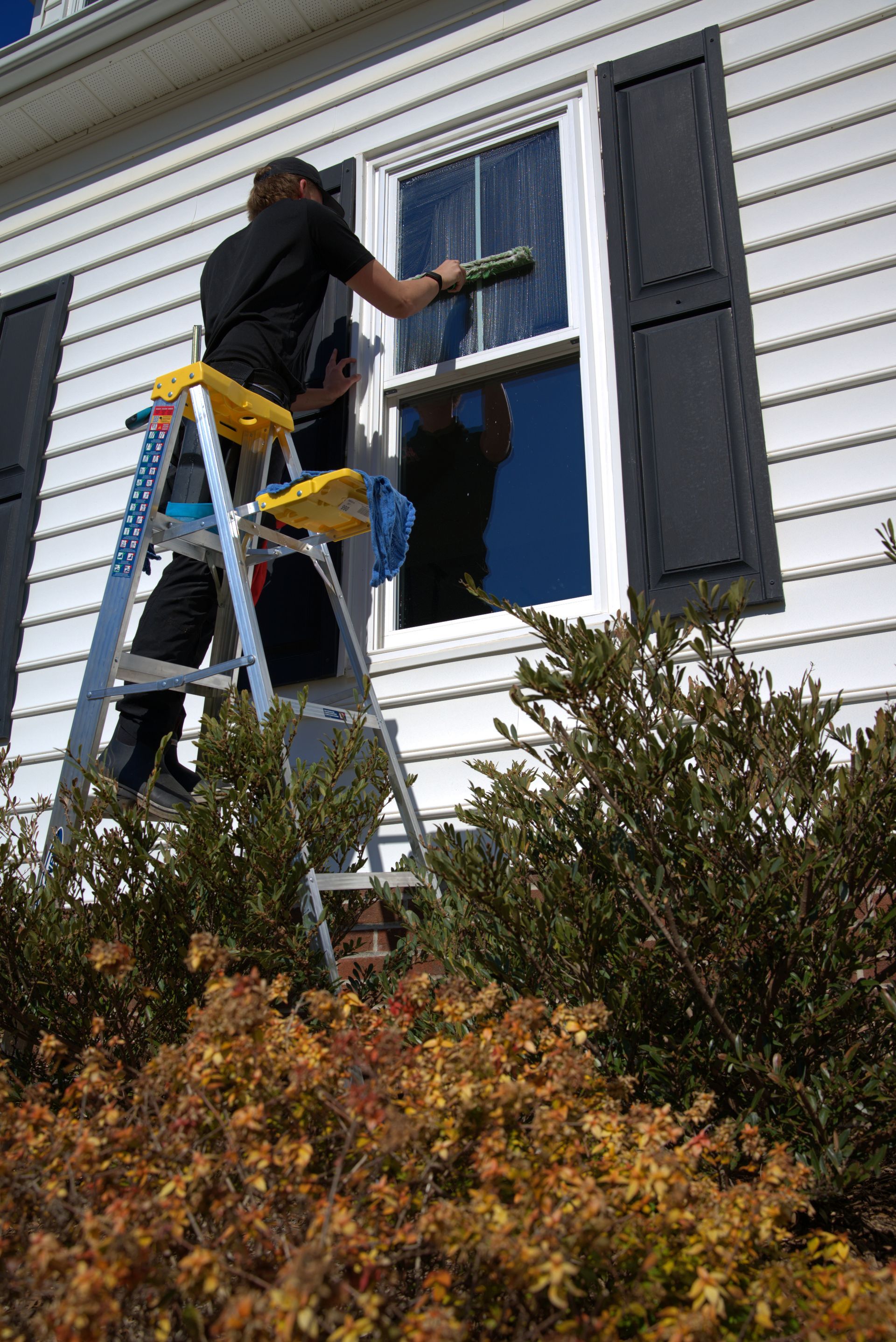 Person cleaning a window on a house exterior using a ladder, squeegee, and bucket.