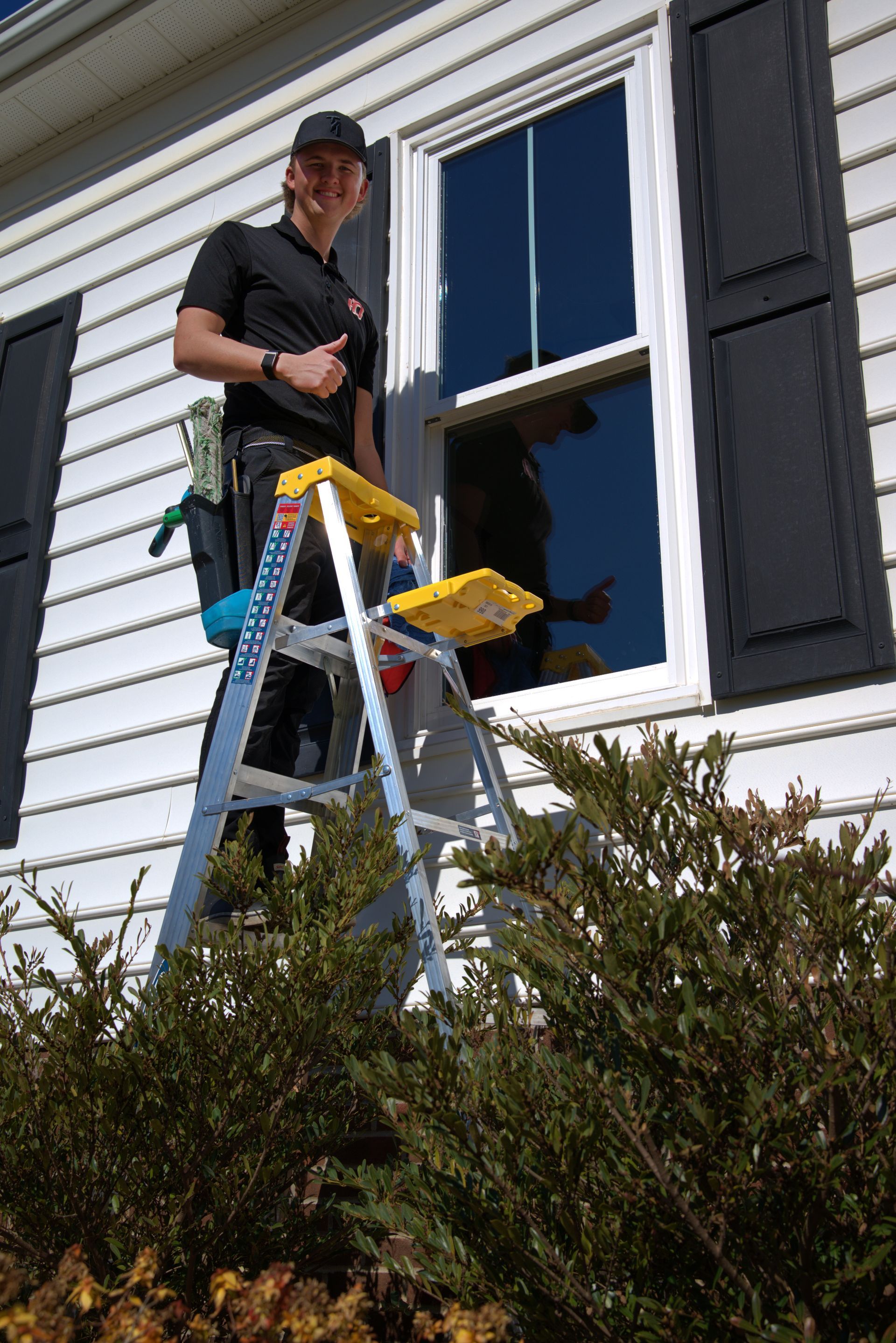 Window washer on ladder gives thumbs up next to house with shutters.