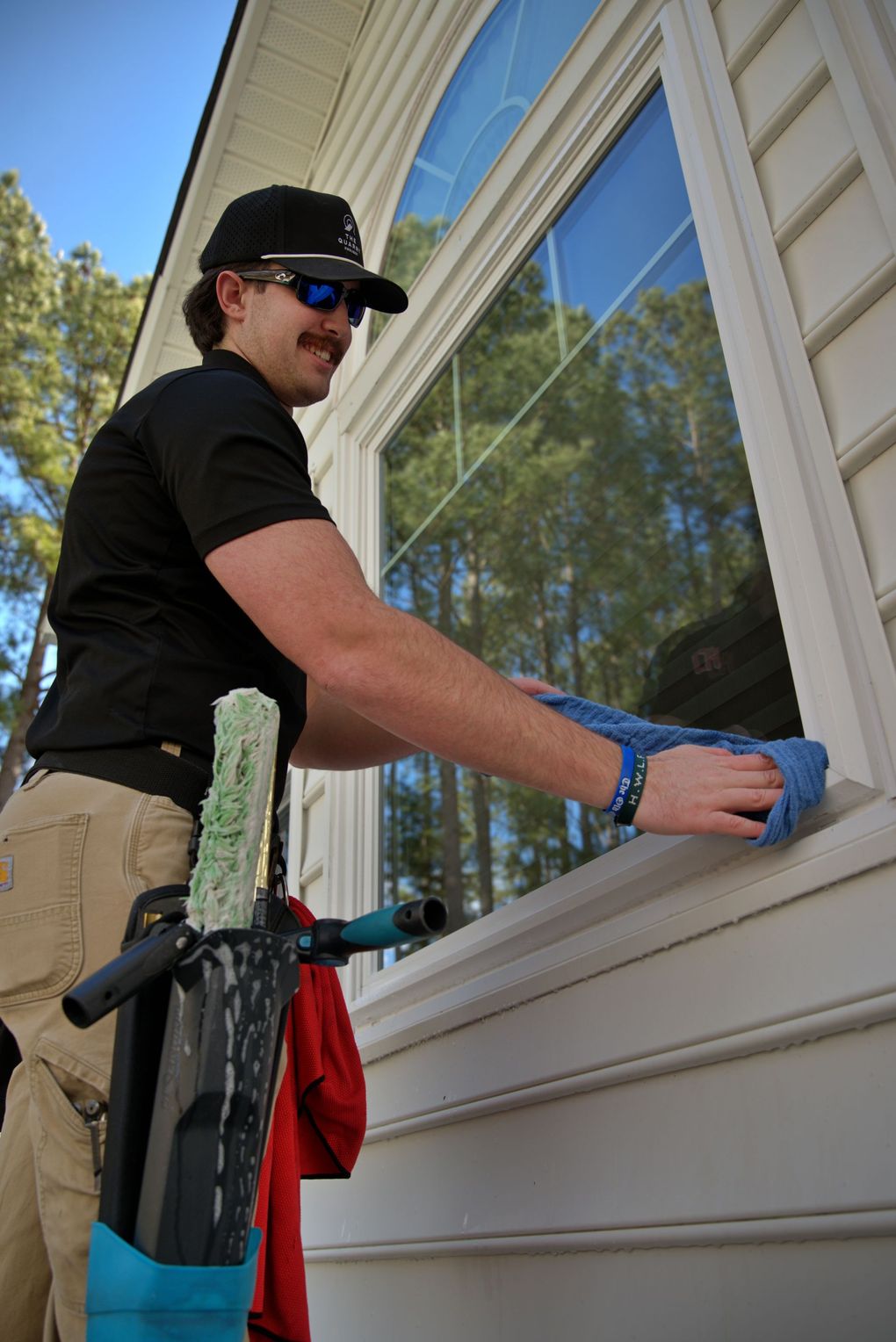 Window cleaner wiping a window outdoors, wearing sunglasses and a baseball cap. A cleaning tool rests below.